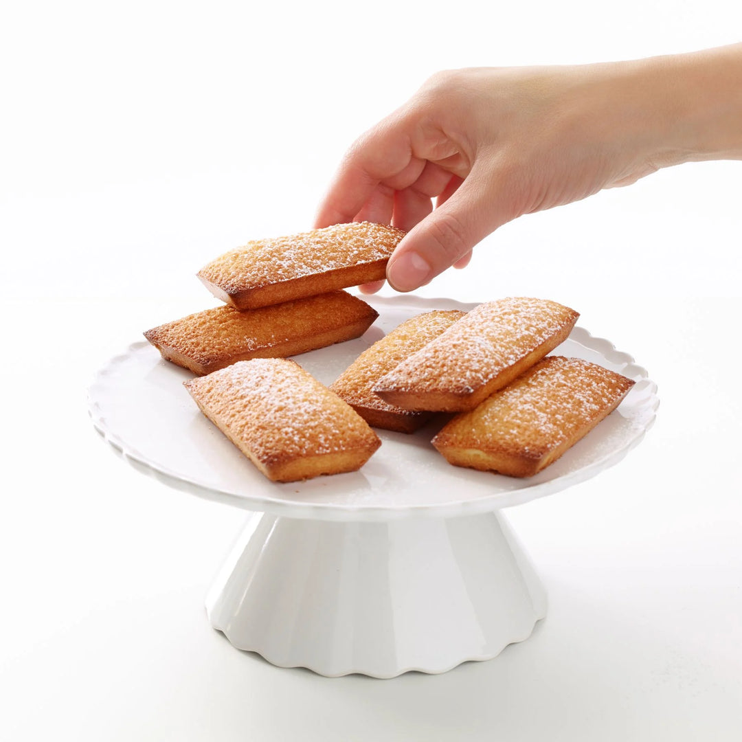 Hand reaching for a madeleine cookie on a white pedestal plate with a plain background