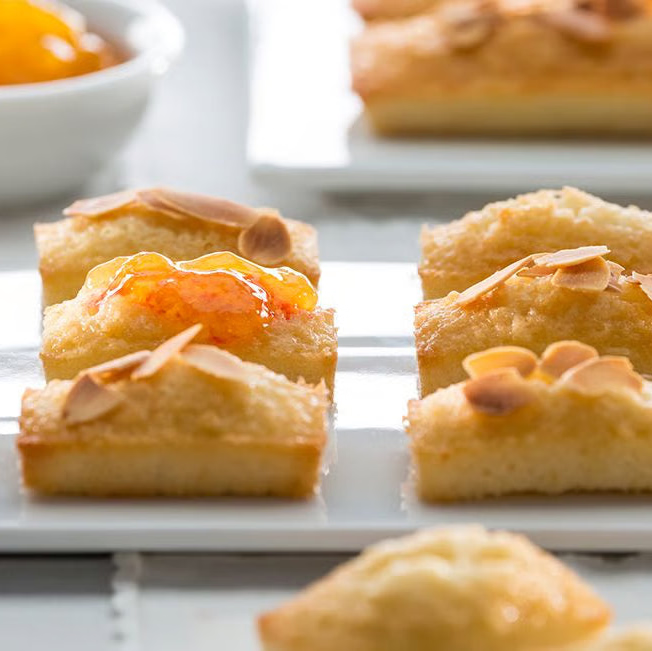 Close-up of several small golden-brown baked pastries topped with sliced almonds and orange jam, arranged on white plates with a small bowl of jam in the background.